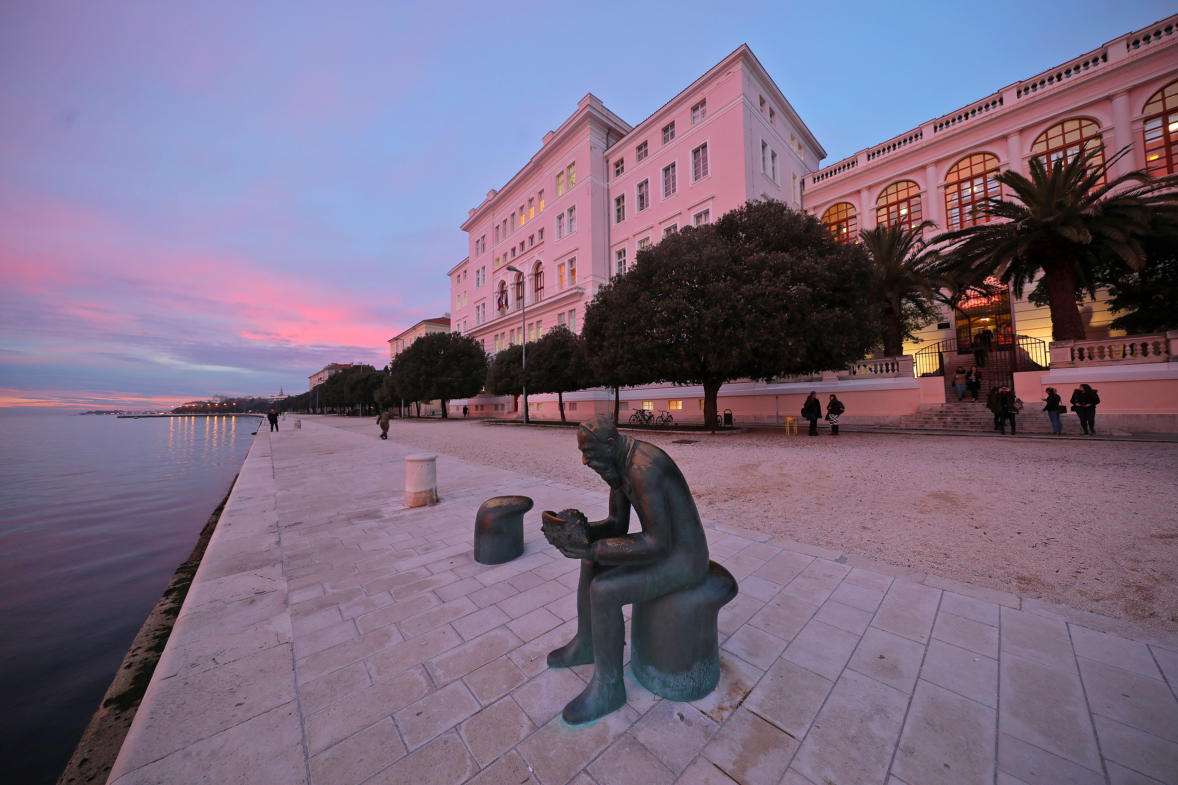 In the foreground, a sculpture of a man sitting down and llooking into a large sea shell. In the background, the University of Zadar at sunset