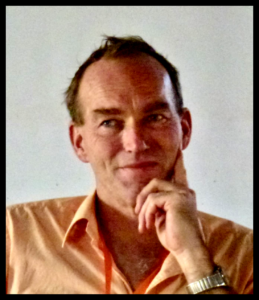 A slightly grain photograph of Thomas Hylland Eriksen, wearing an open-necked, salmon-coloured shirt, with his hand to his chin in a pensive pose